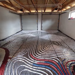 MICHAŁ PIOTROWICZ TECHNIKA GRZEWCZA - Rozłożone białe rurki ogrzewania podłogowego na czarnej siatce w surowym pomieszczeniu z drewnianym stropem i dwoma bramami garażowymi w tle.