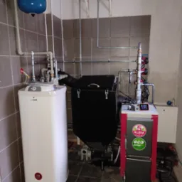 Kompleksowa kotłownia z piecem na paliwo stałe, zasobnikiem CWU, zbiornikiem wyr&oacute;wnawczym i instalacją hydrauliczną w pomieszczeniu z płytkami.