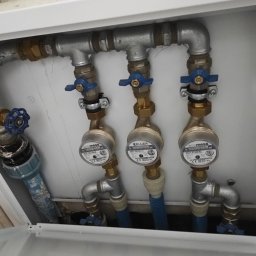 MICHAŁ PIOTROWICZ TECHNIKA GRZEWCZA - Węzeł wodomierzowy z trzema wodomierzami marki Mess i Keller, z niebieskimi zaworami odcinającymi, zamontowany w białej szafce instalacyjnej.