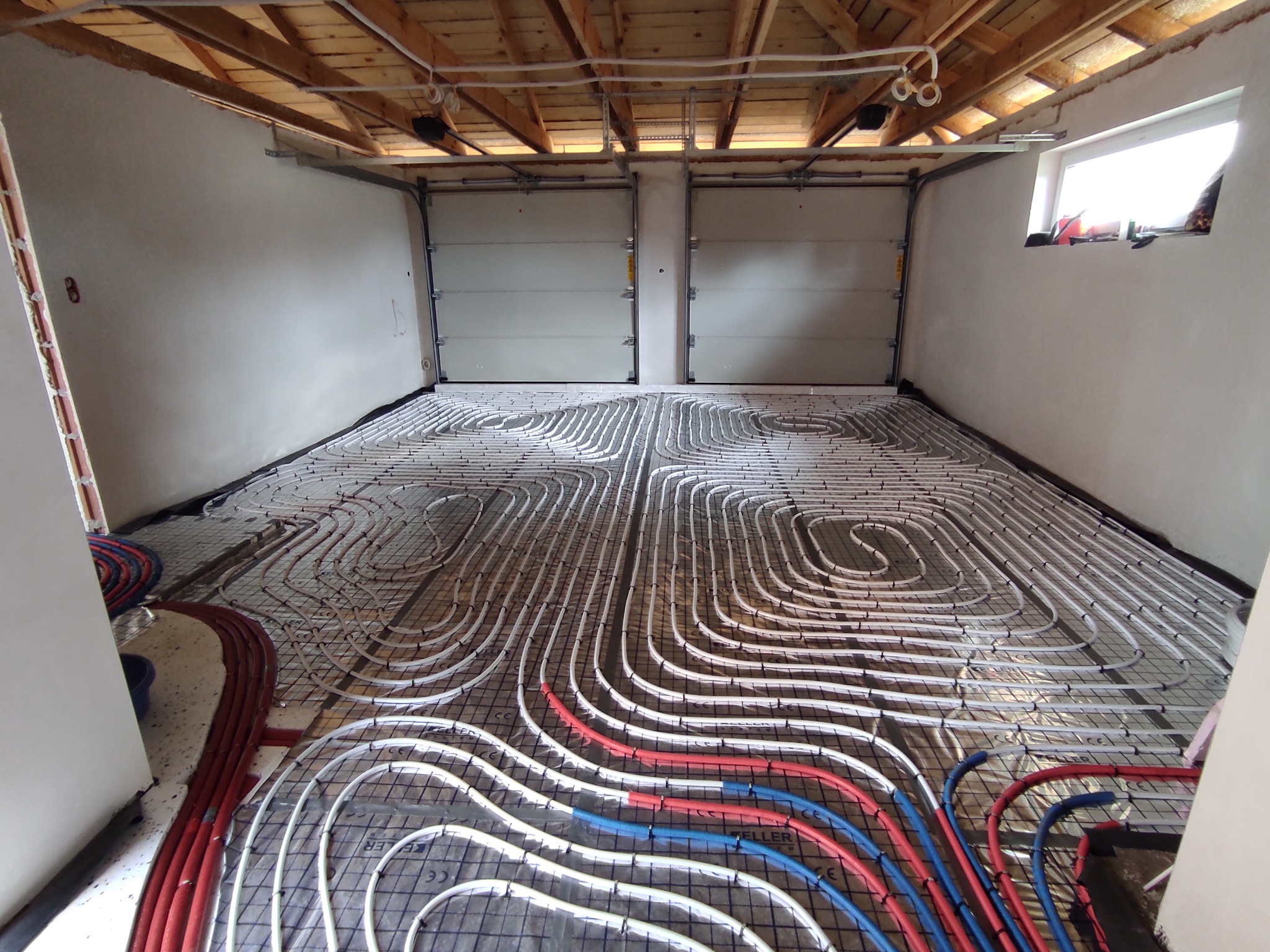Rozłożone białe rurki ogrzewania podłogowego na czarnej siatce w surowym pomieszczeniu z drewnianym stropem i dwoma bramami garażowymi w tle.