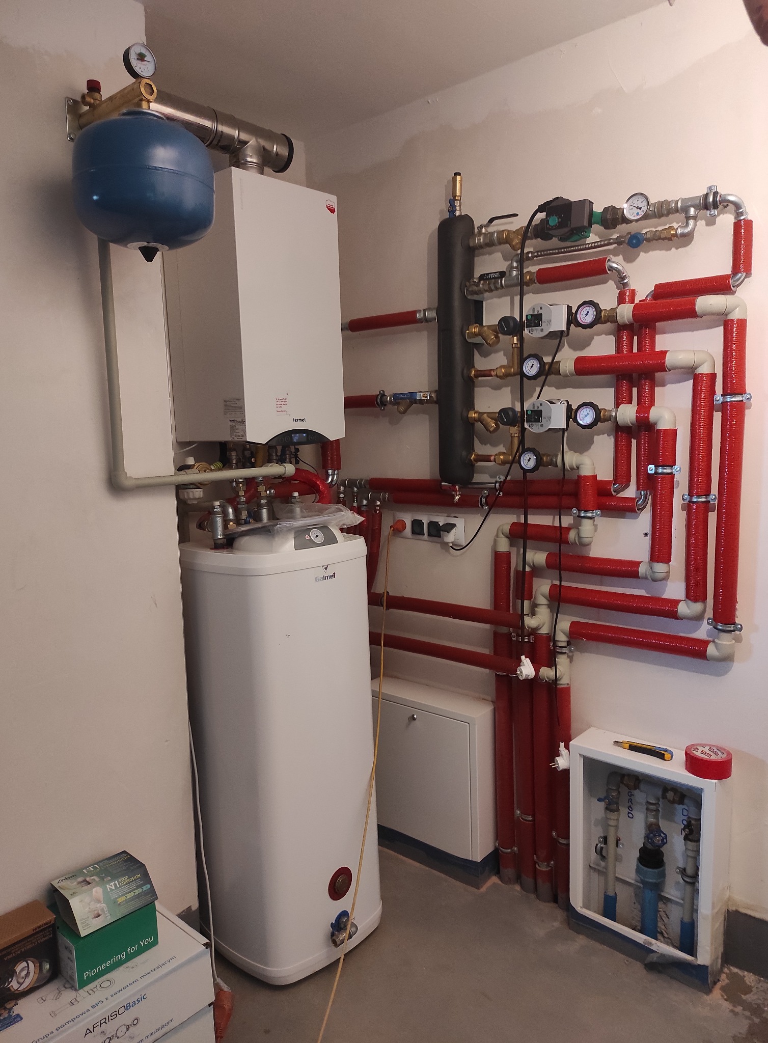 Kompaktowa kotłownia: biały piec gazowy Termet, zasobnik Galmet, niebieski zbiornik wyrównawczy oraz rozbudowany system rur z izolacją termiczną w kolorze czerwonym, manometry i zawory, narzędzia...