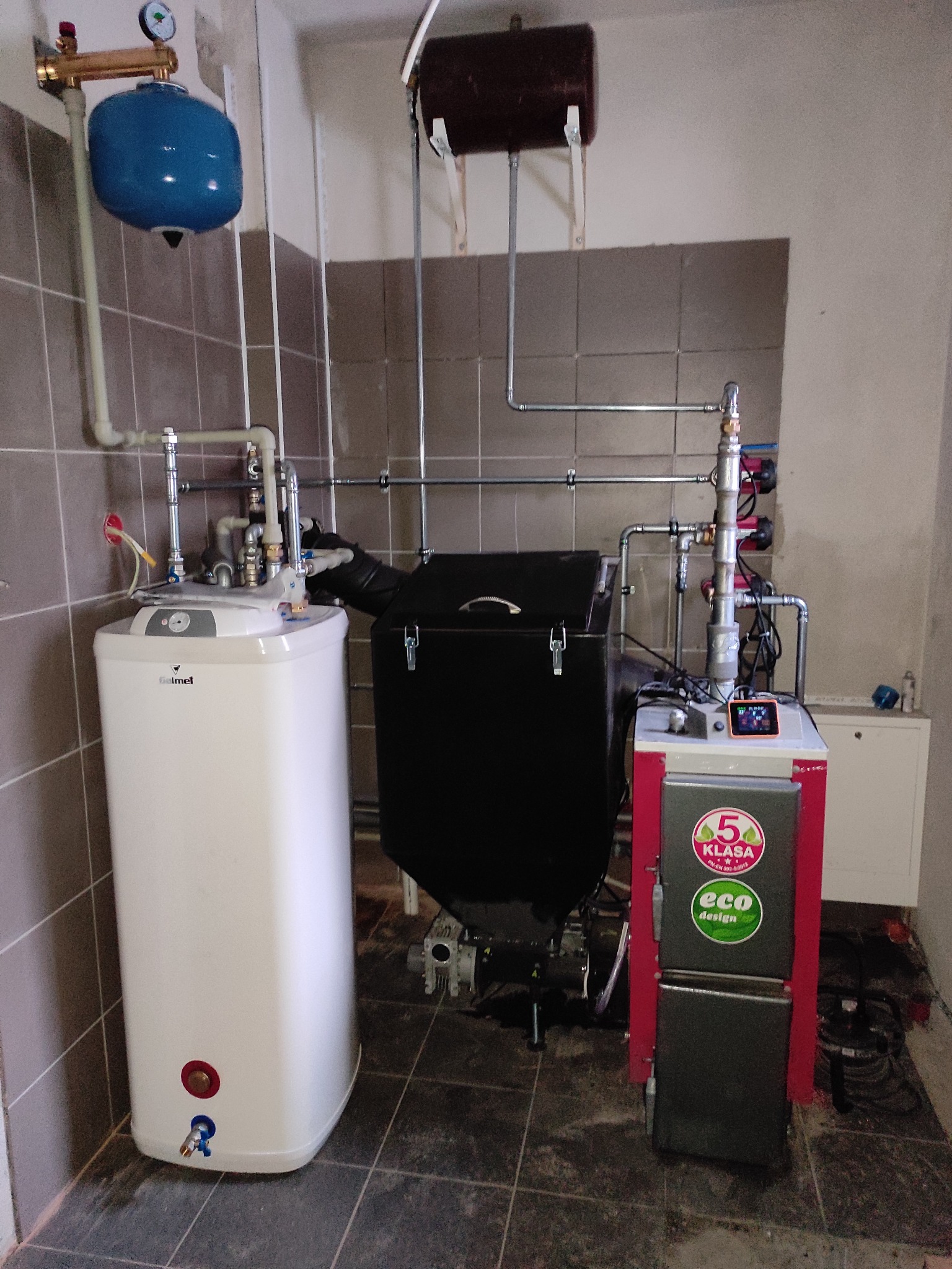 Kompleksowa kotłownia z piecem na paliwo stałe, zasobnikiem CWU, zbiornikiem wyrównawczym i instalacją hydrauliczną w pomieszczeniu z płytkami.