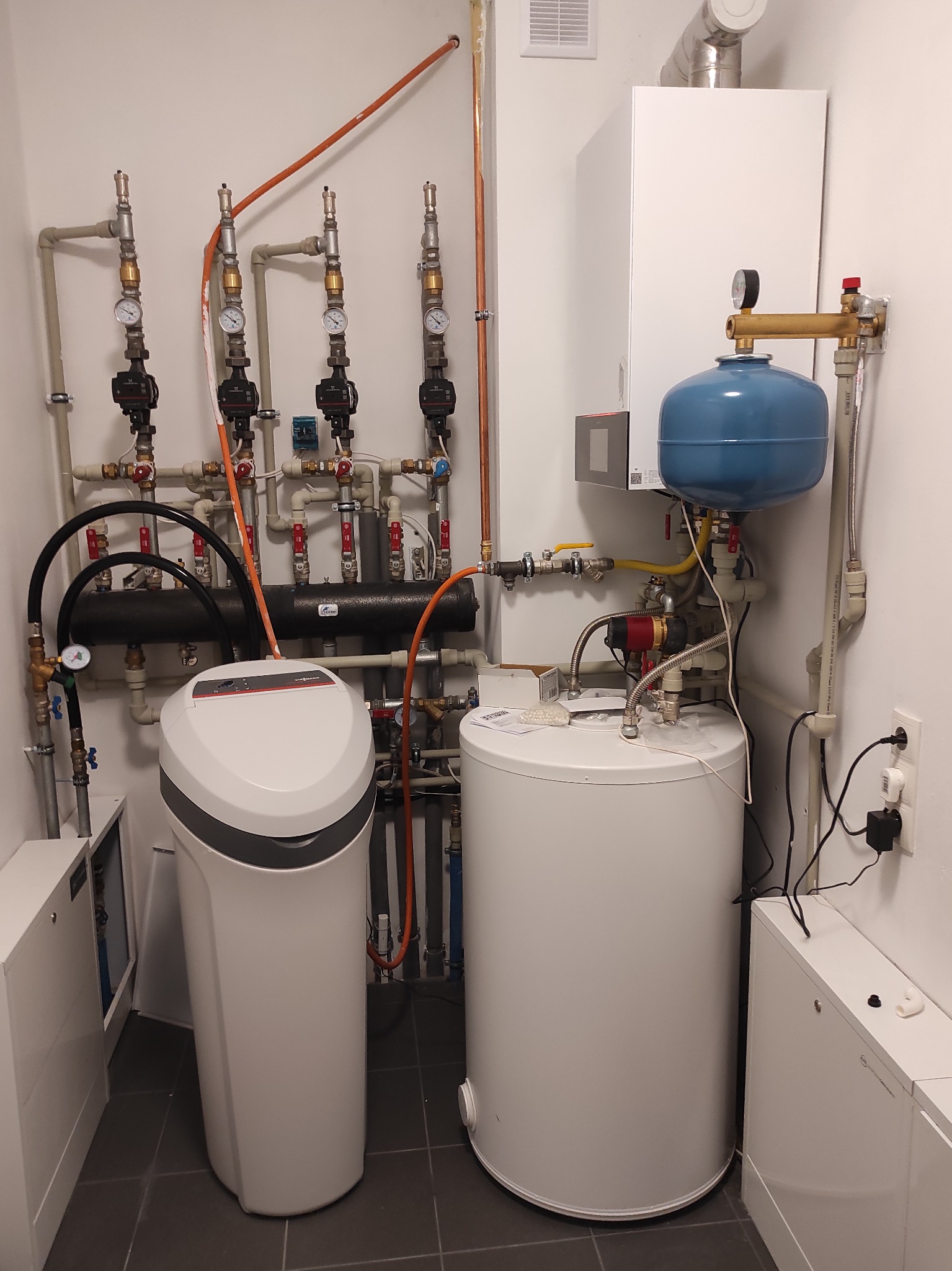 Kotłownia z białym piecem gazowym, niebieskim zbiornikiem wyrównawczym, zmiękczaczem wody Viessmann i rozbudowaną instalacją hydrauliczną z licznymi zaworami i manometrami na szarej płytce podłogowej.