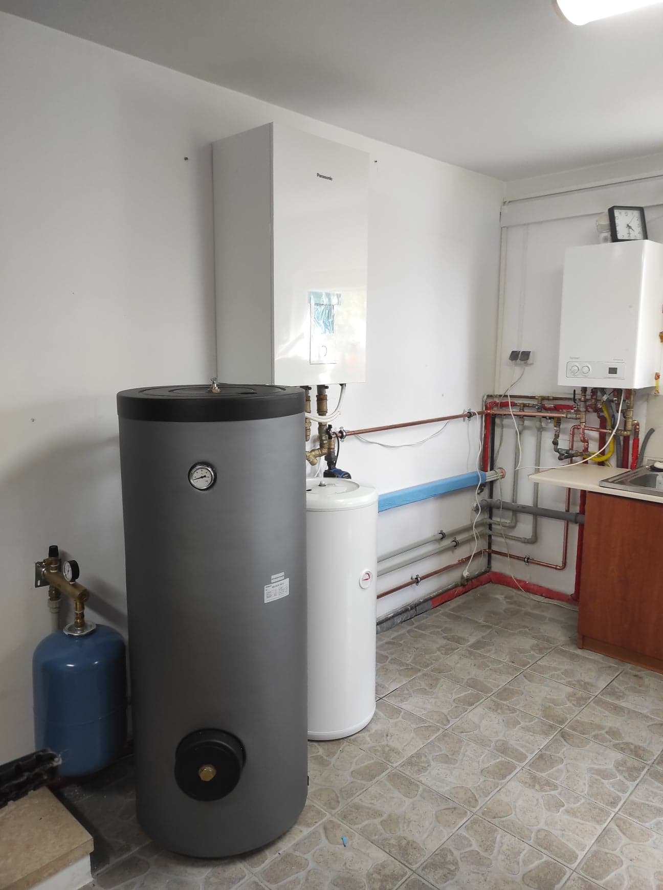 Kotłownia z piecem gazowym Panasonic, zasobnikiem ciepłej wody w kolorze szarym i białym, oraz niebieskim naczyniem przeponowym. Widoczne rury miedziane i instalacja hydrauliczna na ścianie.
