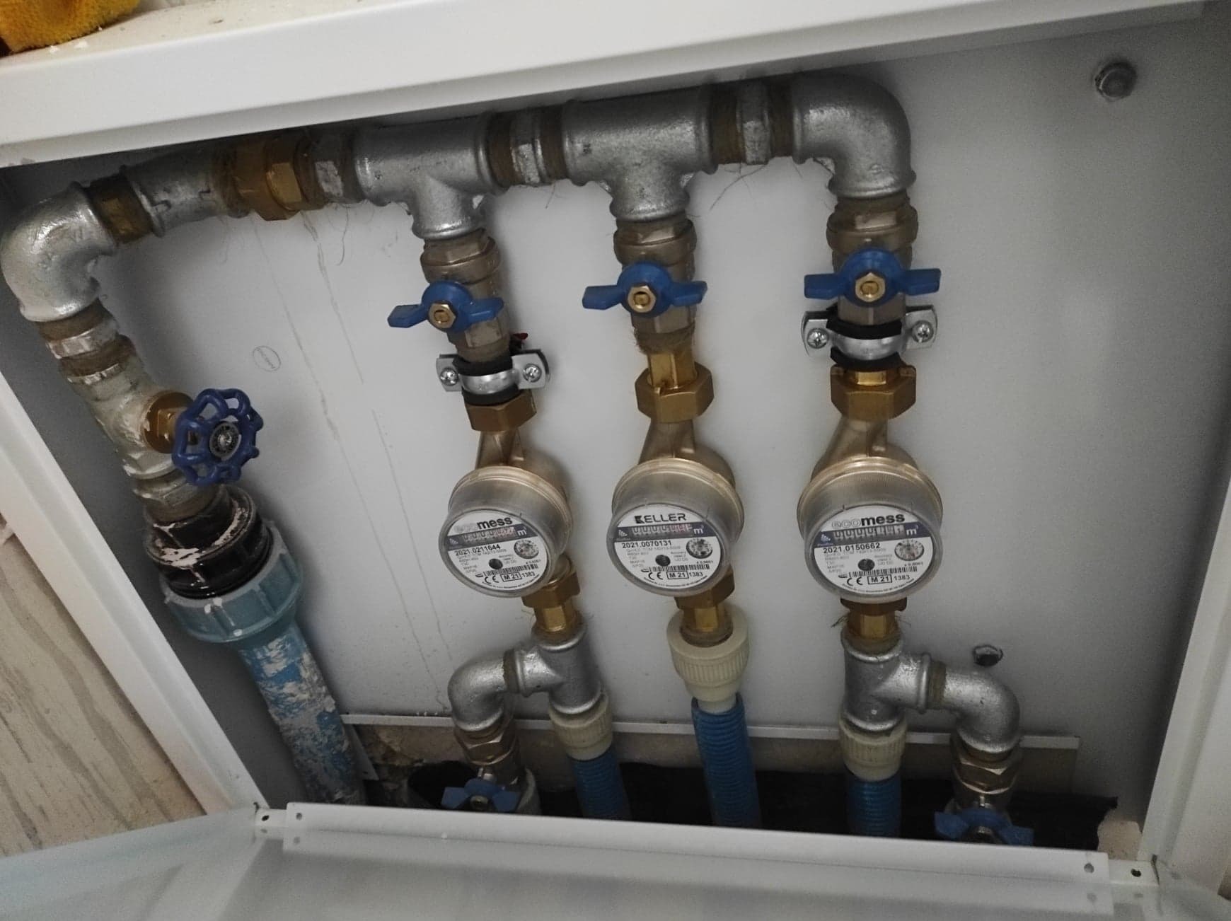 Węzeł wodomierzowy z trzema wodomierzami marki Mess i Keller, z niebieskimi zaworami odcinającymi, zamontowany w białej szafce instalacyjnej.