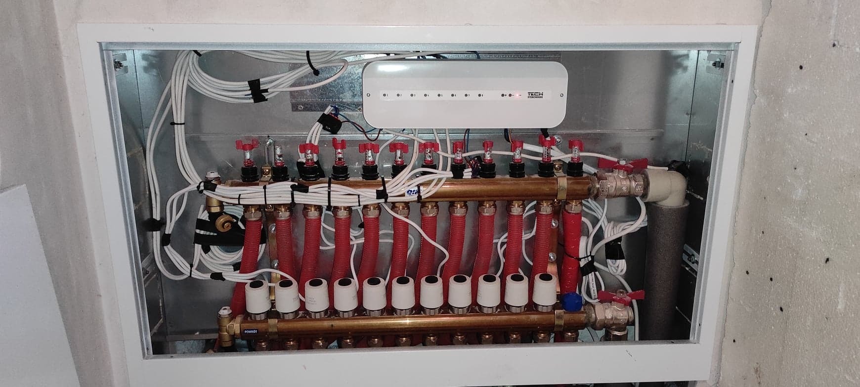 Rozdzielacz ogrzewania podłogowego w metalowej szafce z licznymi czerwonymi rurami, białymi przewodami i sterownikiem TECH, zamontowany na surowej ścianie.