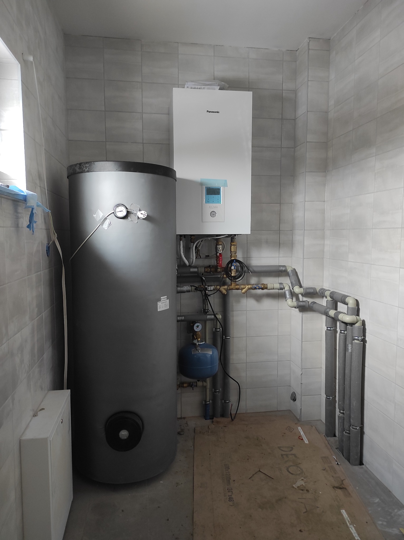Kotłownia z zamontowanym piecem gazowym Panasonic i zasobnikiem ciepłej wody, widoczne rury instalacji grzewczej z izolacją termiczną, manometry zabezpieczone taśmą.
