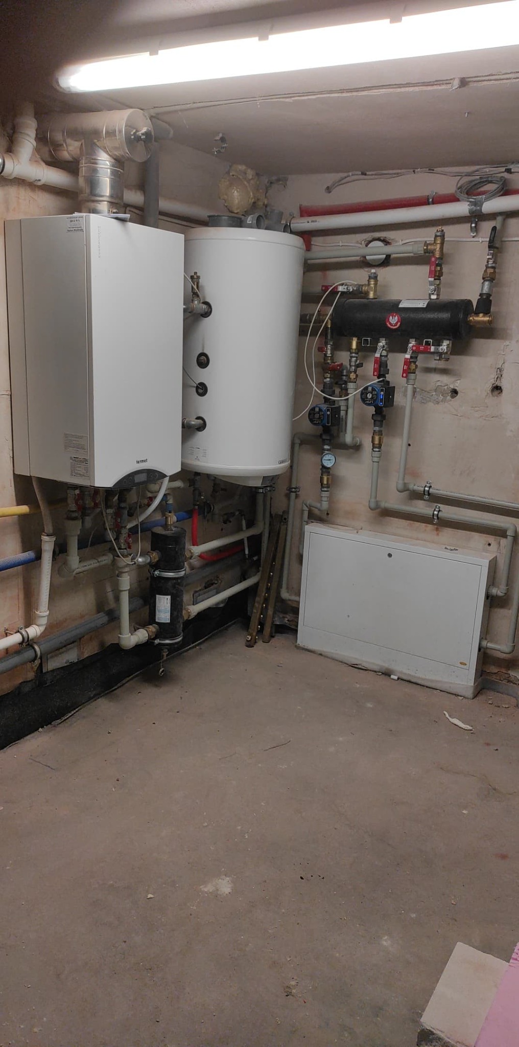 Kocioł gazowy Termet wiszący na ścianie obok białego zasobnika CWU, z widocznymi rurami i osprzętem w piwnicy.