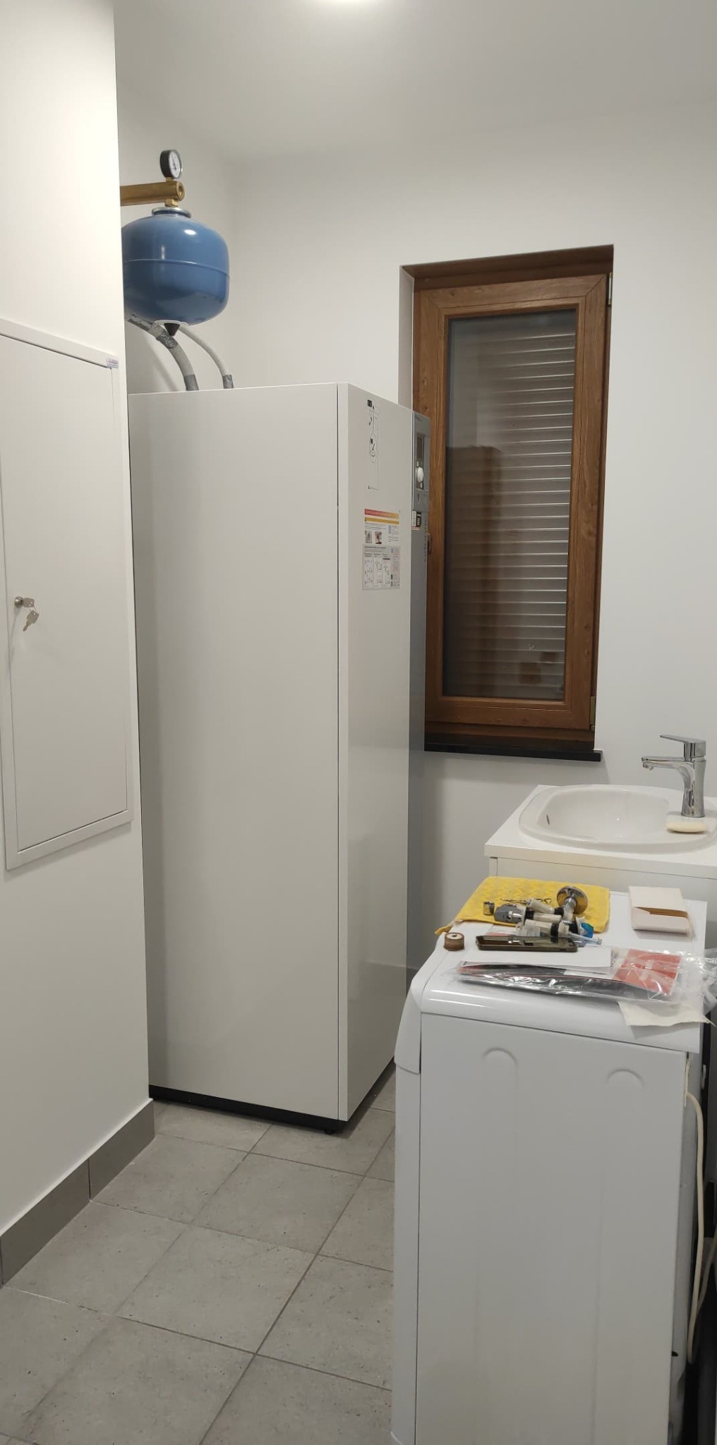 Biały piec grzewczy z niebieskim zbiornikiem wyrównawczym w jasnym pomieszczeniu, widoczne narzędzia hydrauliczne na pralce, okno z drewnianą ramą w tle.