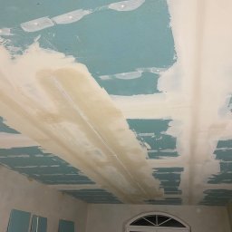 BRYNBUD Marian Bryn - Niedokończony sufit z płyt kartonowo-gipsowych, widoczne szpachlowane łączenia, surowa powierzchnia, ślady niedokładnego szpachlowania, niebieska farba bazowa.