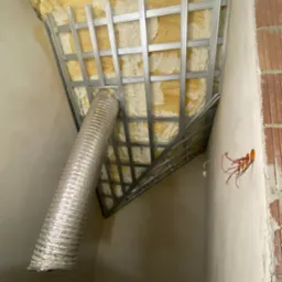 Sufit poddasza w trakcie izolacji: widoczna metalowa konstrukcja pod płyty, pianka izolacyjna oraz wystający aluminiowy przewód wentylacyjny, surowe ściany z instalacją elektryczną.