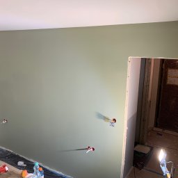Oleksandr Kirichenko - Ściana pomalowana na oliwkowy kolor z widocznymi puszkami elektrycznymi. Podłoga zabezpieczona folią malarską. Widoczne narzędzia malarskie i karton.