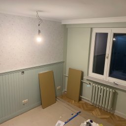 Oleksandr Kirichenko - Pokój w trakcie remontu: pomalowane ściany do połowy na zielono, tapeta w górnej części, grzejnik, wisząca żarówka i panele czekające na montaż.