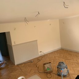 Oleksandr Kirichenko - Jasne wnętrze w trakcie remontu: parkiet w jodełkę, ściany w kremowym odcieniu, widoczne przewody elektryczne i narzędzia. Fachowiec w tle.