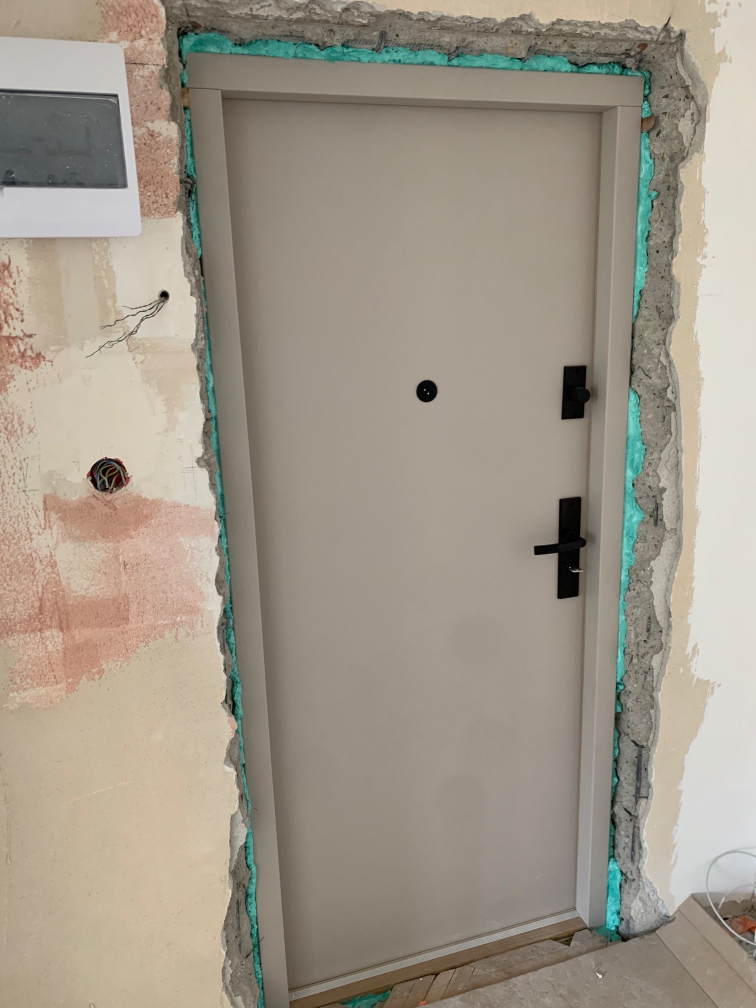 Montaż beżowych drzwi wejściowych z czarną klamką. Ościeżnica uszczelniona pianką montażową, ściana w stanie surowym z widocznymi instalacjami.