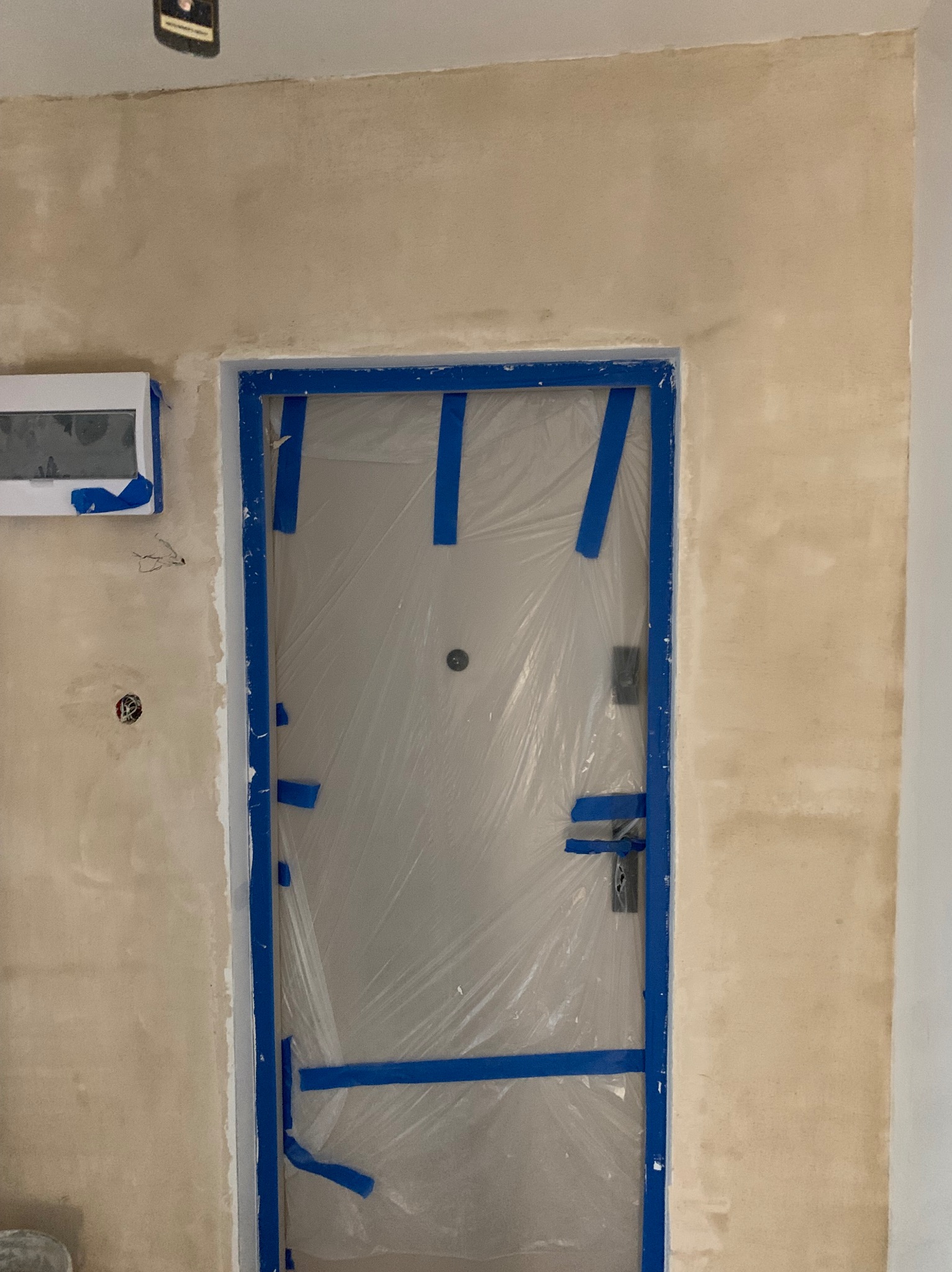 Przygotowanie do malowania: zabezpieczone folią i taśmą drzwi wejściowe, ściana po gładzi, gotowa do malowania. Widoczna puszka elektryczna i klimatyzator.