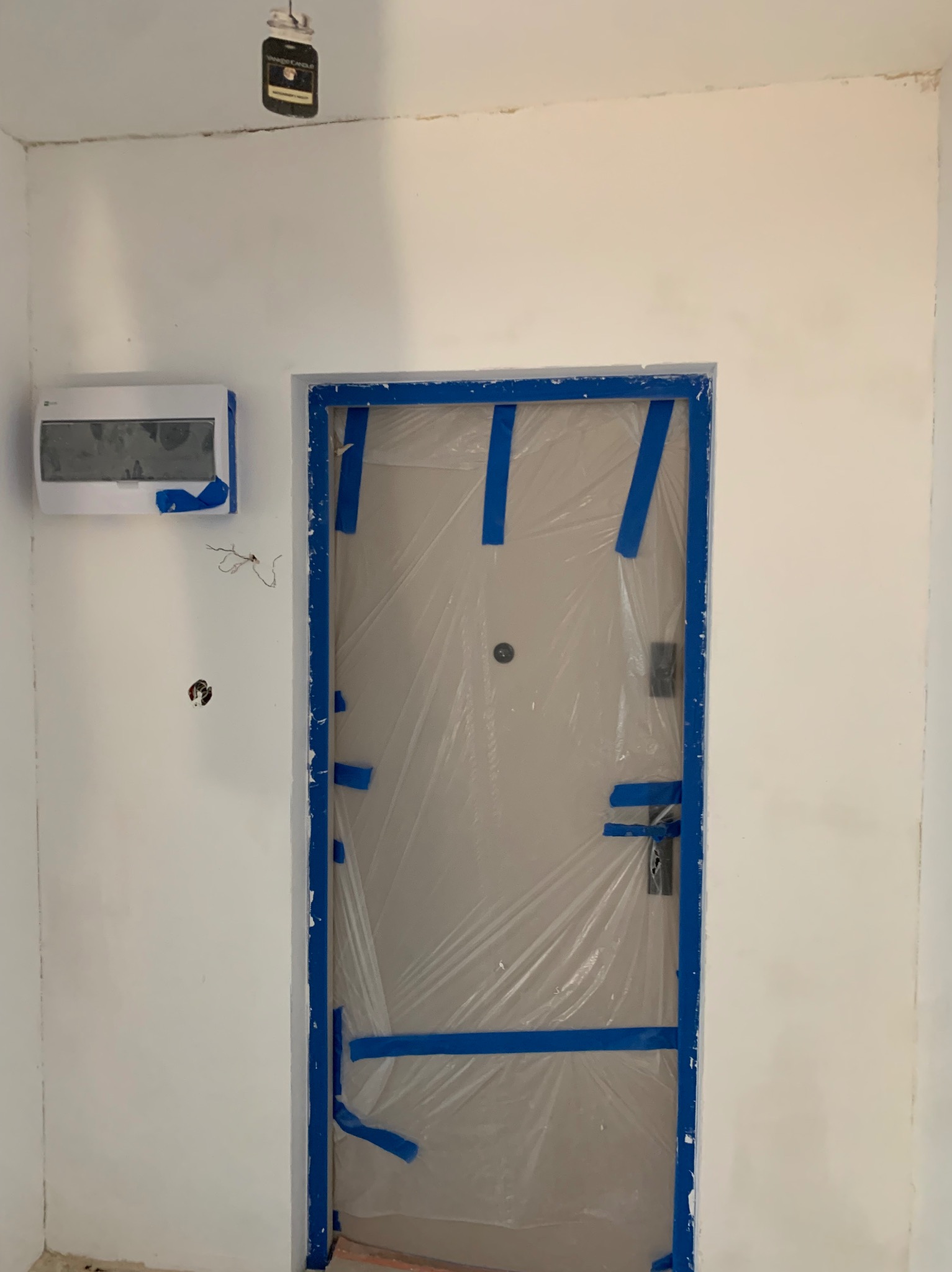 Przygotowanie do malowania: zabezpieczone folią i taśmą drzwi w białym wnętrzu. Widoczna puszka elektryczna i klimatyzator, również zabezpieczone taśmą malarską.