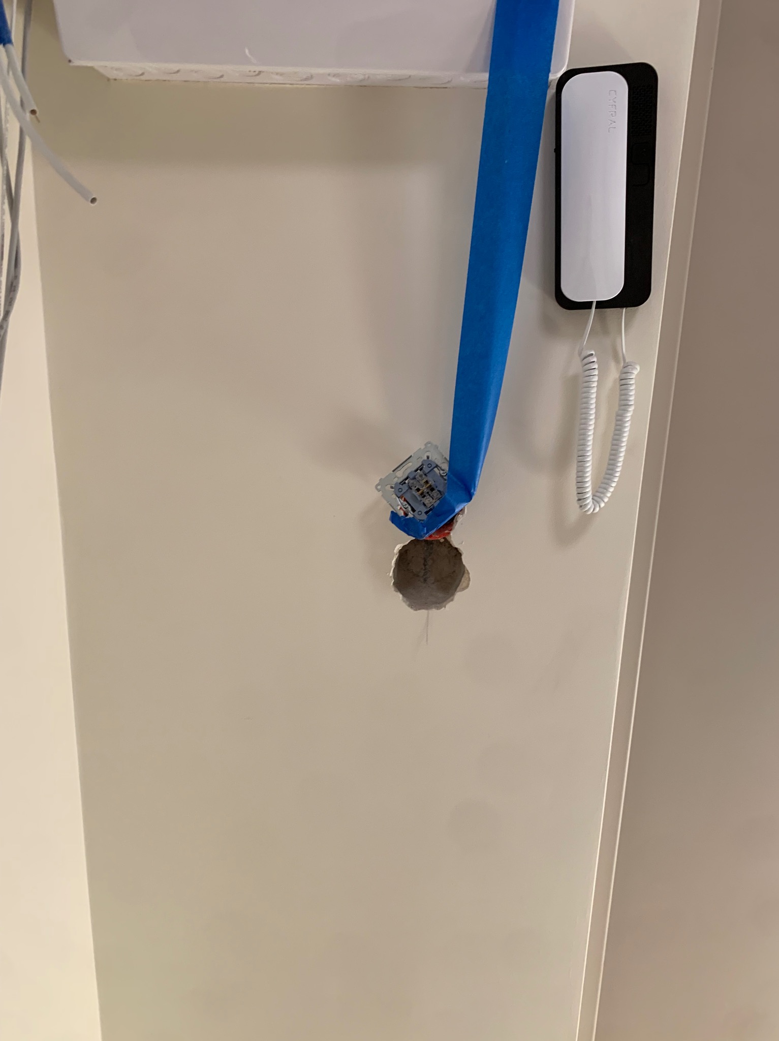 Montaż gniazdka elektrycznego w ścianie, zabezpieczonego taśmą malarską. Widoczny interkom. Prace remontowe w trakcie, surowy otwór w ścianie.