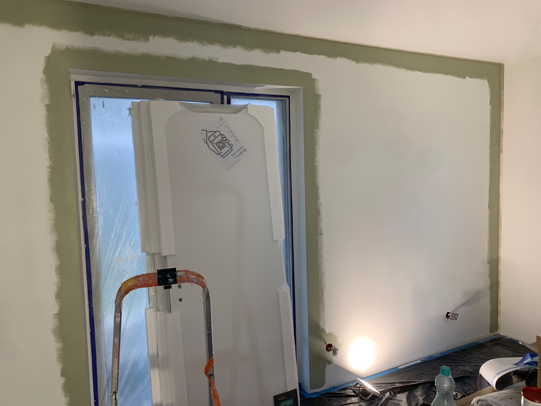 Ściana w trakcie malowania z widocznymi odcięciami w kolorze oliwkowym, zabezpieczone okno folią i drabina malarska. Etap przygotowania do malowania.