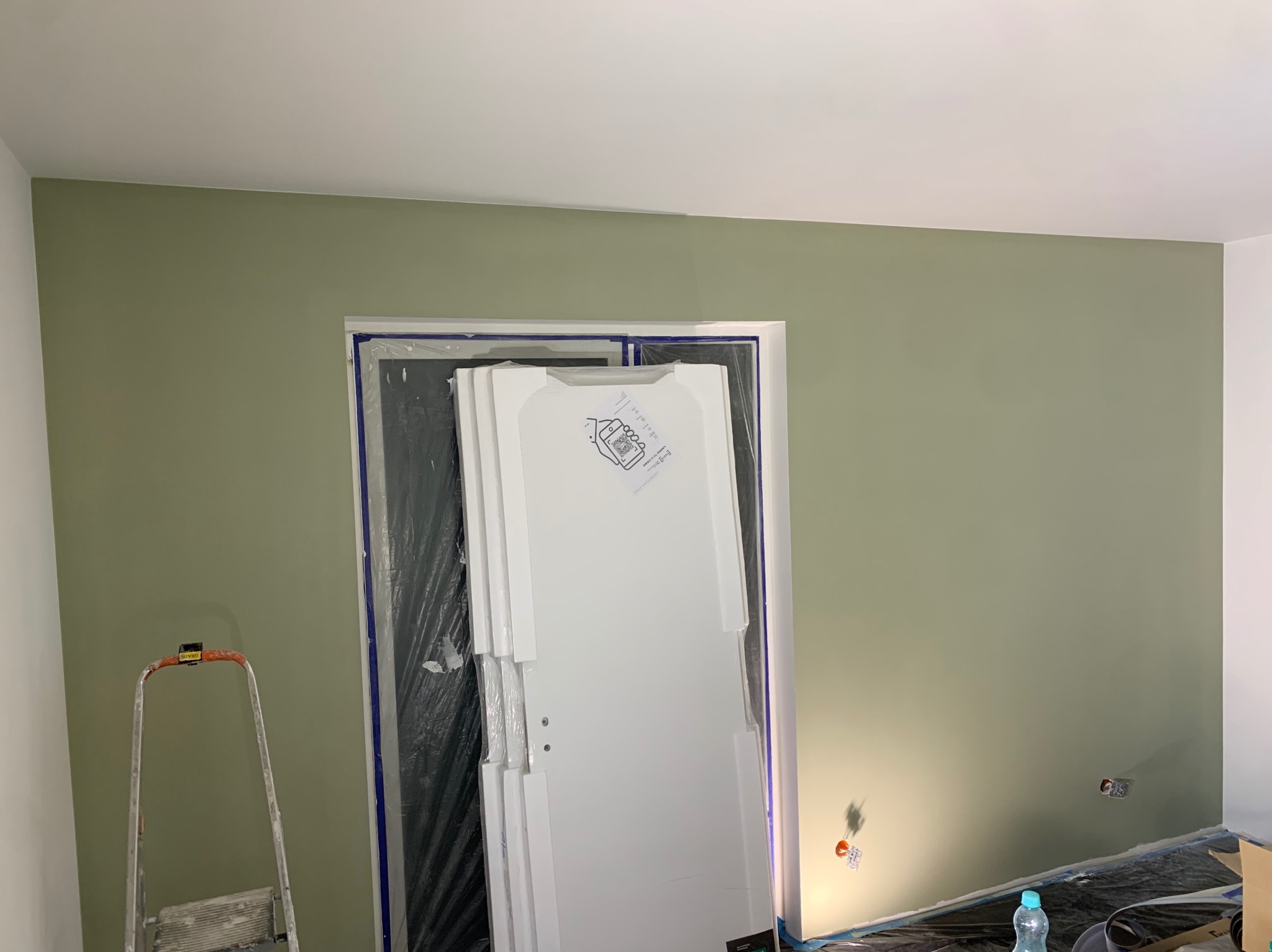 Ściana pomalowana na oliwkowy kolor, zabezpieczone drzwi folią, drabina malarska. Widoczne gniazdka elektryczne. Etap prac wykończeniowych w mieszkaniu.