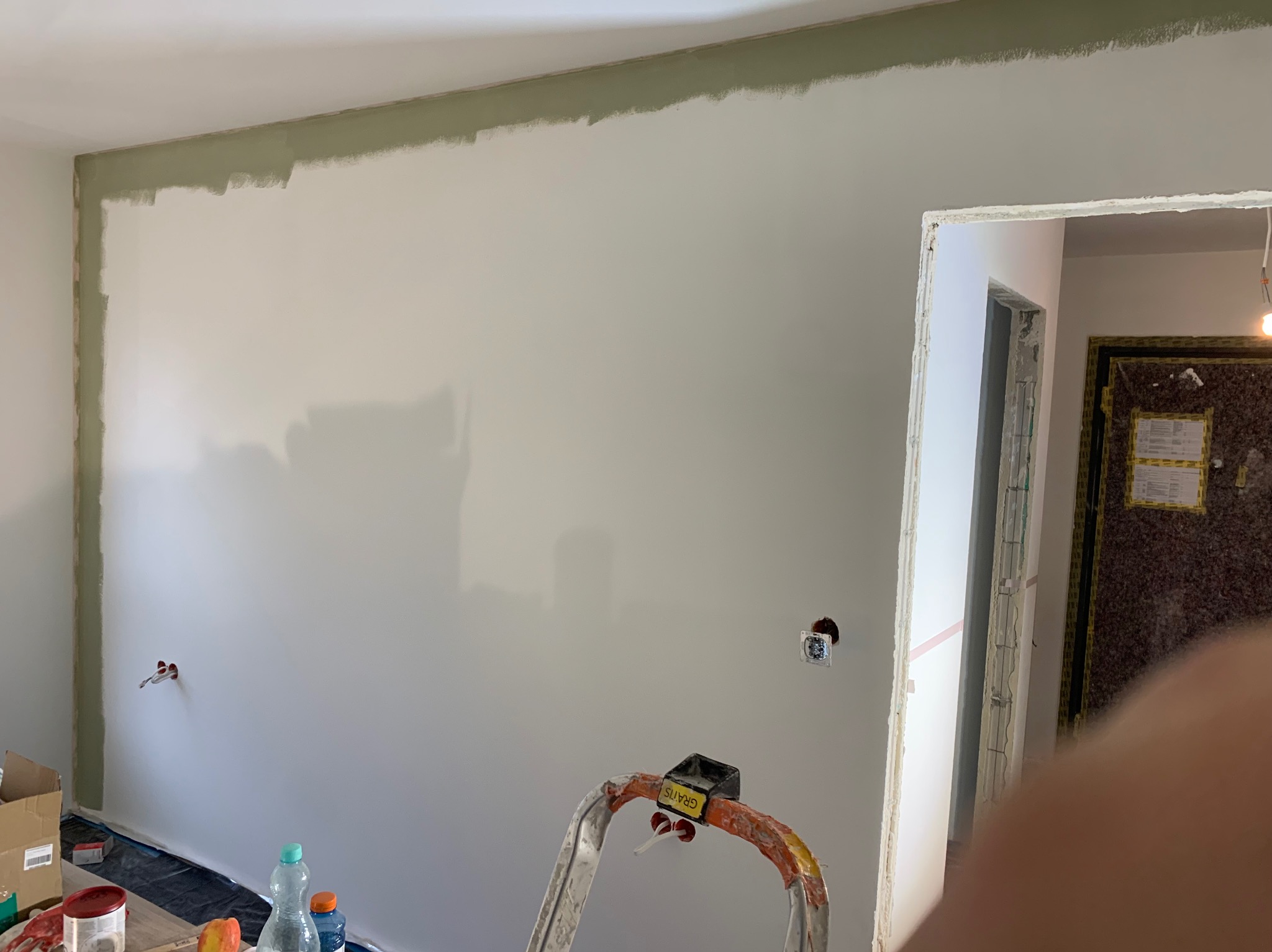 Ściana w trakcie malowania z widoczną obwódką w kolorze oliwkowym, drabina malarska i zabezpieczona folią podłoga. Widoczne gniazdka elektryczne.