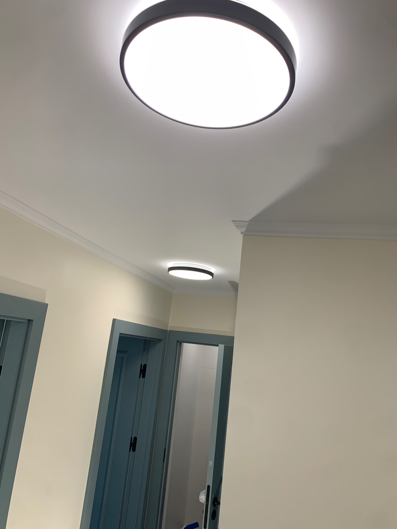 Nowoczesne oświetlenie sufitowe LED w jasnym korytarzu z kremowymi ścianami i niebieskimi futrynami drzwi. Dwa okrągłe plafony równomiernie oświetlają przestrzeń.