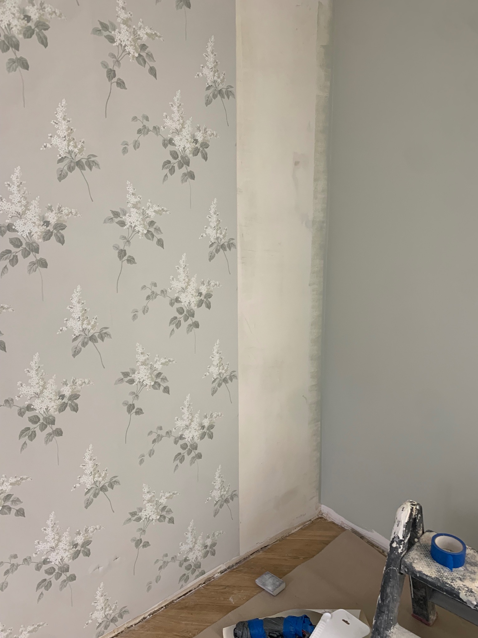 Ściana po częściowym tapetowaniu w kwiatowy wzór, obok fragment ściany pomalowanej na jednolity, jasny kolor. Widoczne narzędzia i drabina malarska.