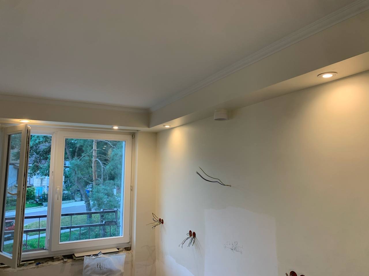 Jasne wnętrze po remoncie: świeżo pomalowane ściany w odcieniach bieli, okno z widokiem na zieleń, widoczne punkty oświetleniowe i przygotowane instalacje elektryczne.