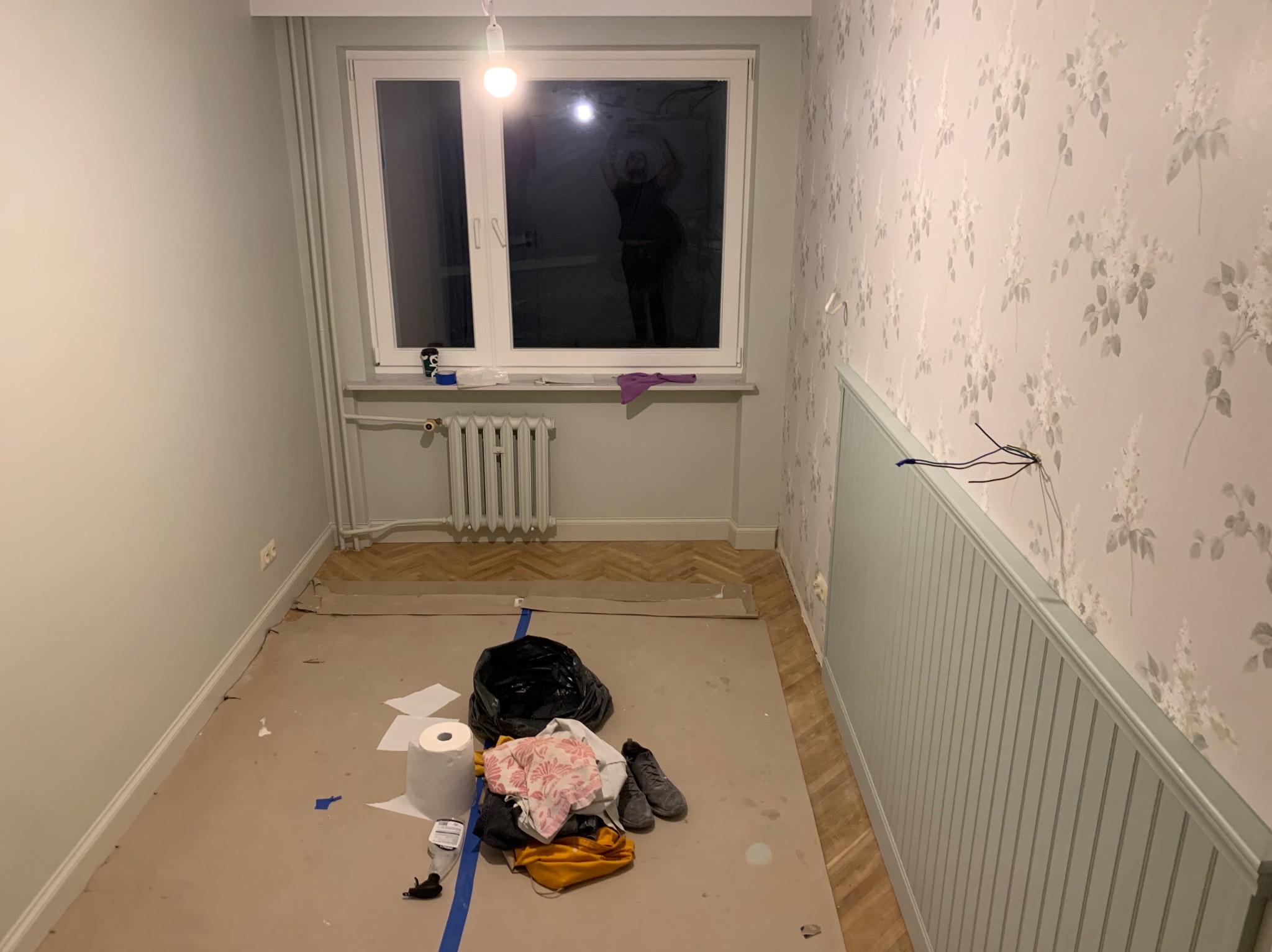 Pokój w trakcie remontu: ściany pomalowane na szaro, jedna ściana z tapetą w kwiaty, widoczne narzędzia i materiały malarskie, okno z odbiciem osoby.