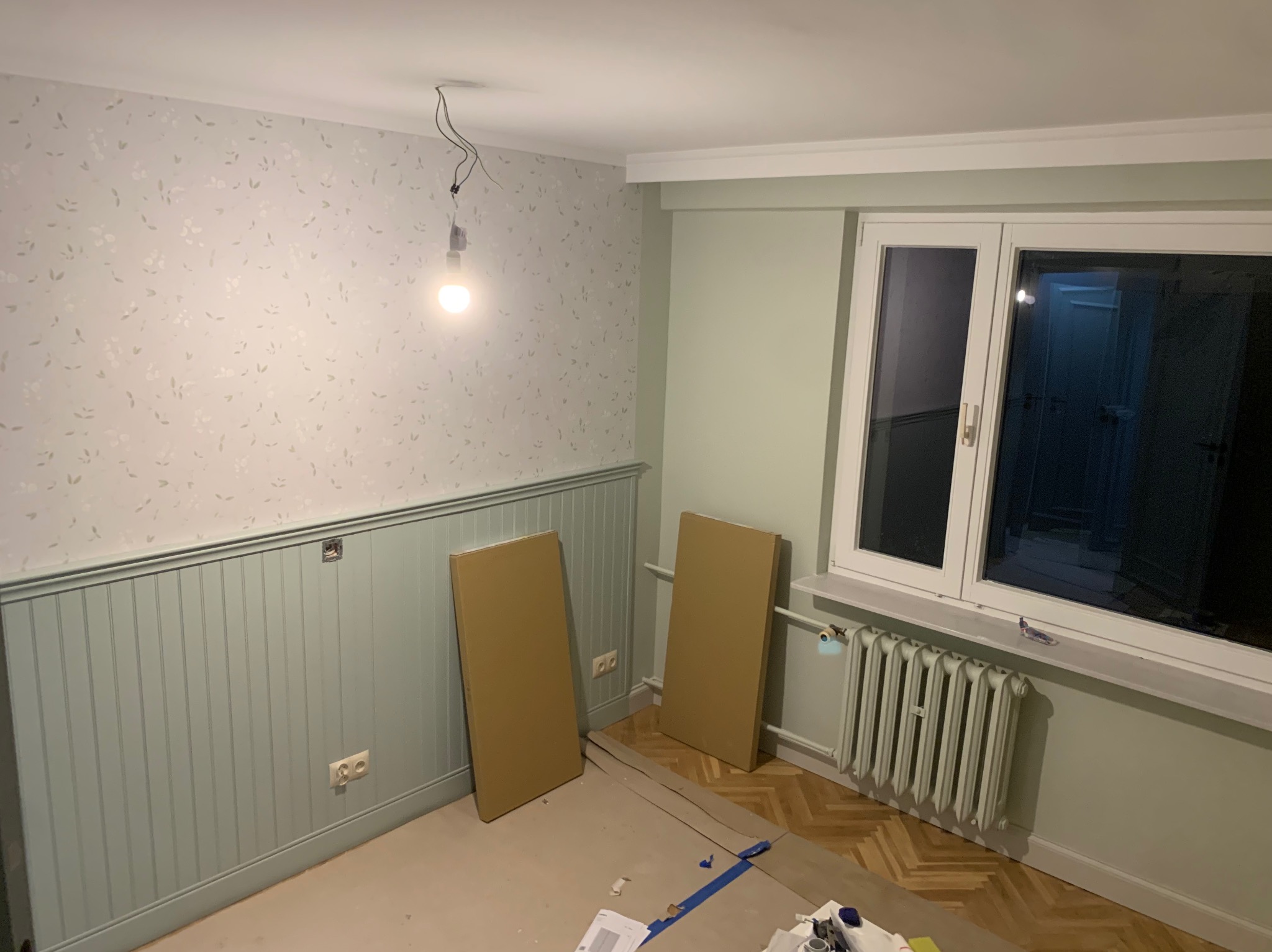 Pokój w trakcie remontu: pomalowane ściany do połowy na zielono, tapeta w górnej części, grzejnik, wisząca żarówka i panele czekające na montaż.
