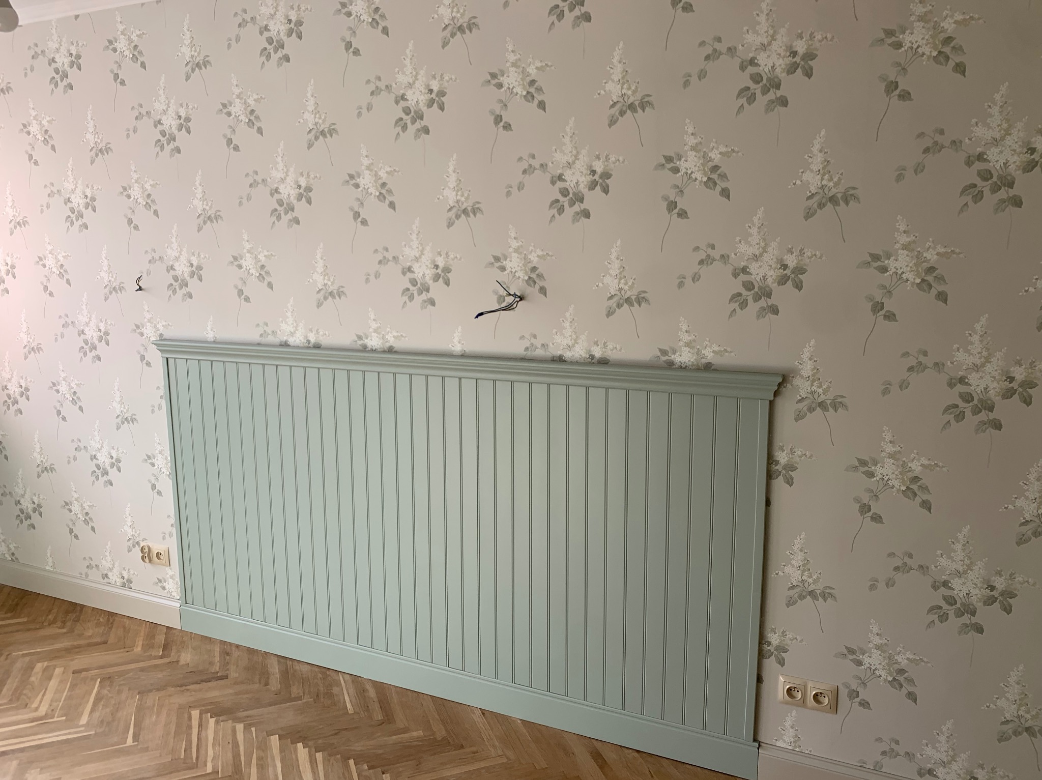 Ściana z tapetą w kwiaty i panelami ściennymi w odcieniu zieleni, parkiet w jodełkę. Widoczne gniazdka elektryczne. Dekoracyjne wykończenie wnętrza.