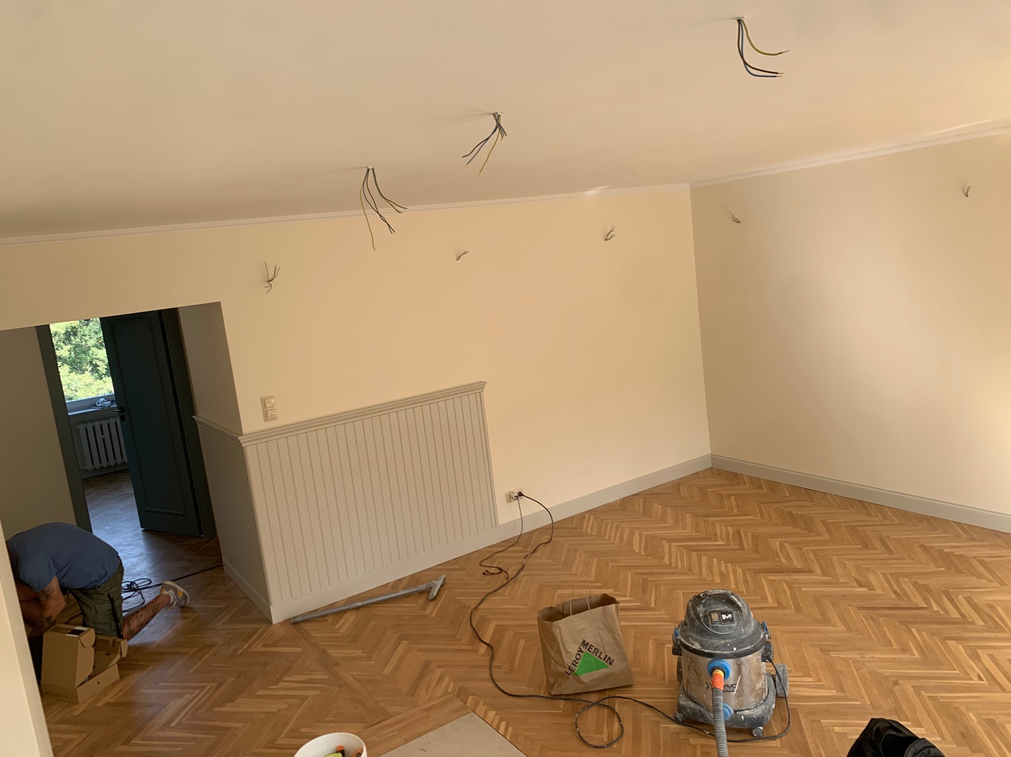 Jasne wnętrze w trakcie remontu: parkiet w jodełkę, ściany w kremowym odcieniu, widoczne przewody elektryczne i narzędzia. Fachowiec w tle.