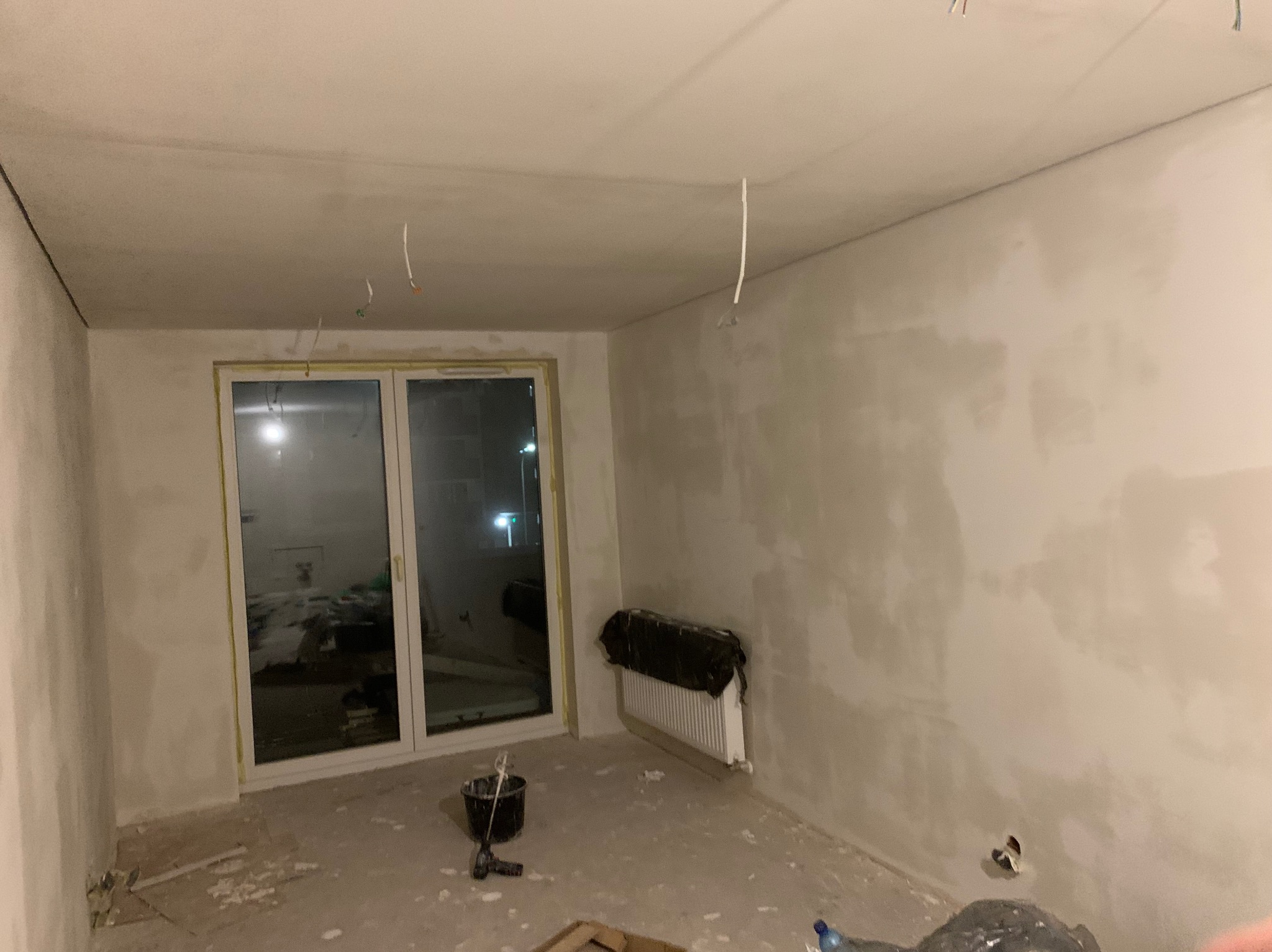 Pokój po gładziowaniu ścian, widoczne narzędzia malarskie, okno balkonowe i zakryty grzejnik. Przygotowanie do malowania ścian w nowym mieszkaniu.