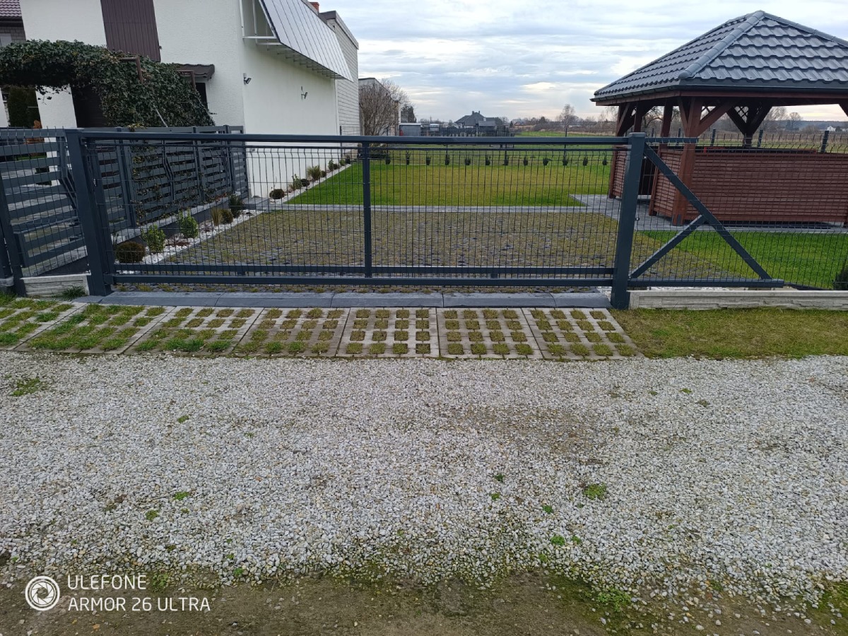 Przesuwna brama wjazdowa z metalową siatką na posesję z kostką ażurową i szarym żwirem, w tle altana i dom.