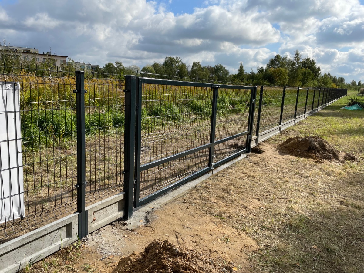 Długa linia ogrodzenia z siatki z bramą wjazdową, osadzona na betonowej podmurówce, widok z zewnątrz posesji, świeżo usypana ziemia przy fundamencie.