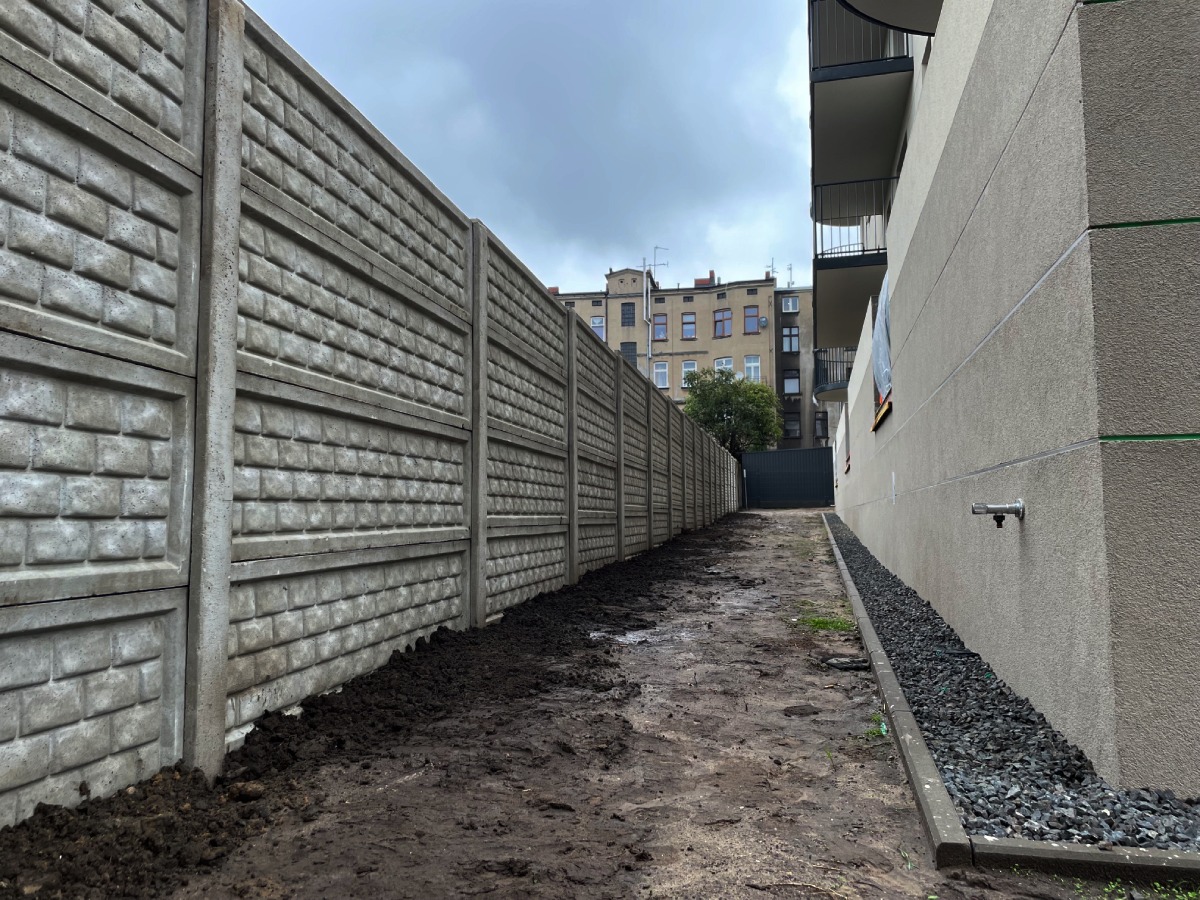 Długi, szary mur z prefabrykatów betonowych imitujących cegłę, oddzielający budynek od drogi, z widoczną ziemią i fragmentem budynku w tle.