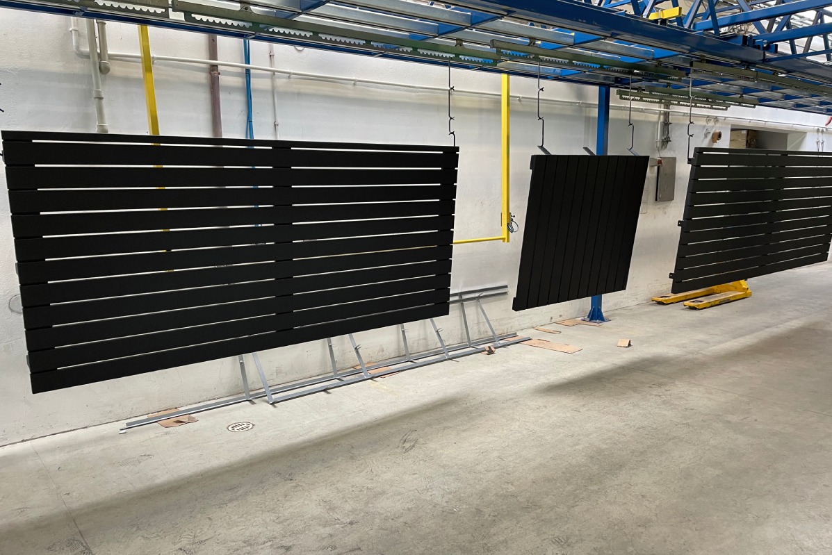 Czarne, poziome elementy ogrodzenia na linii produkcyjnej, zawieszone na hakach, widoczne metalowe podpory i paleciak.