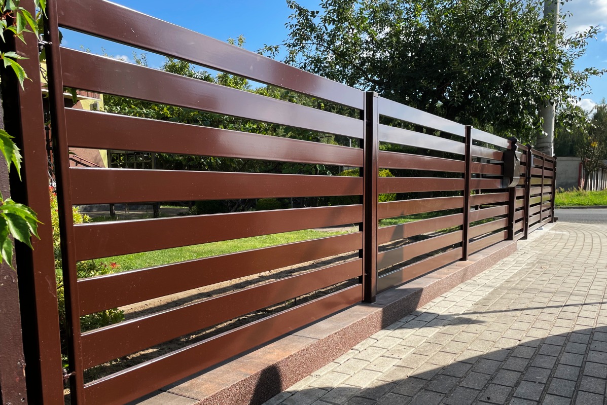 Nowoczesne ogrodzenie panelowe z poziomych profili w kolorze brązowym, zamontowane na niskiej podmurówce z kostki, widok z chodnika.