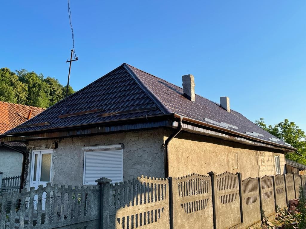 Dom z nowym, brązowym dachem, rynny zamontowane, widoczne kominy i antena. Betonowe ogrodzenie na pierwszym planie. Słoneczny dzień.