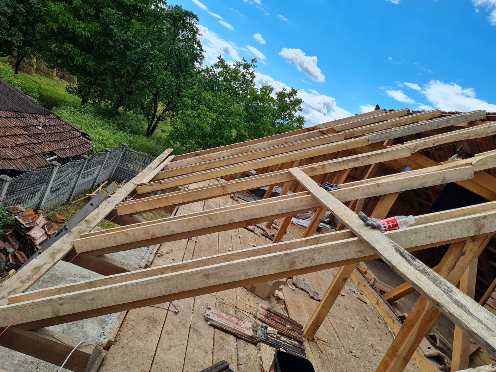 Drewniana konstrukcja dachu w trakcie wymiany, widoczne krokwie i łaty na tle błękitnego nieba z chmurami. Częściowo zdjęte stare dachówki. Prace dekarskie w toku.