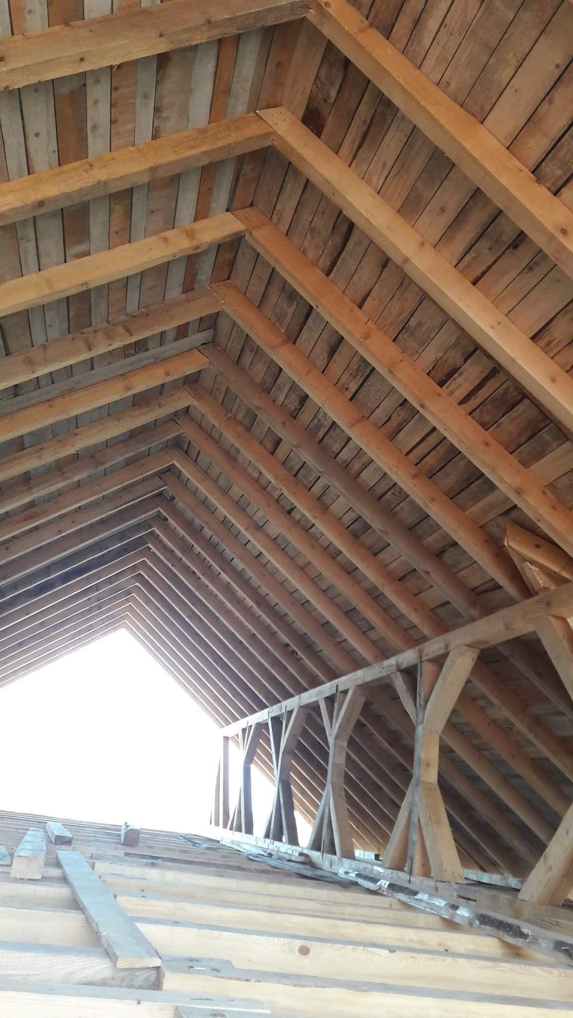 Drewniana konstrukcja dachu z widocznymi krokwiami i deskowaniem, w trakcie budowy lub remontu. Widok od wewnątrz, z prześwitem światła dziennego.