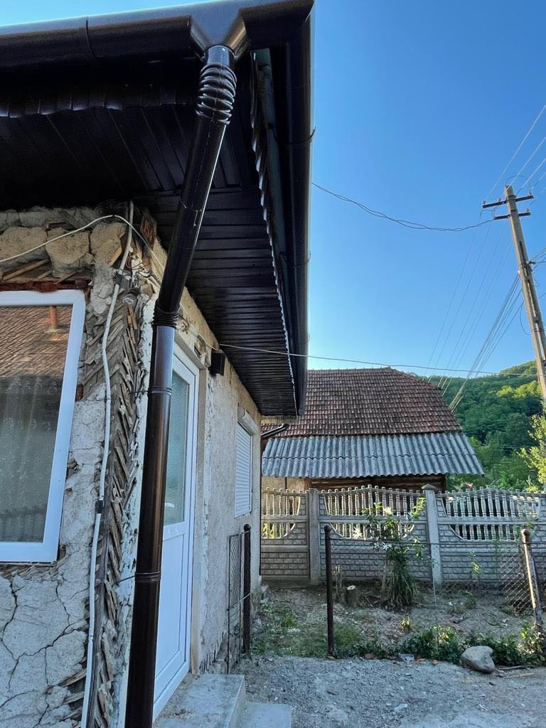 Ciemnobrązowa rynna spustowa zamontowana na budynku z kamienną elewacją. W tle widoczny dach drugiego budynku i błękitne niebo.