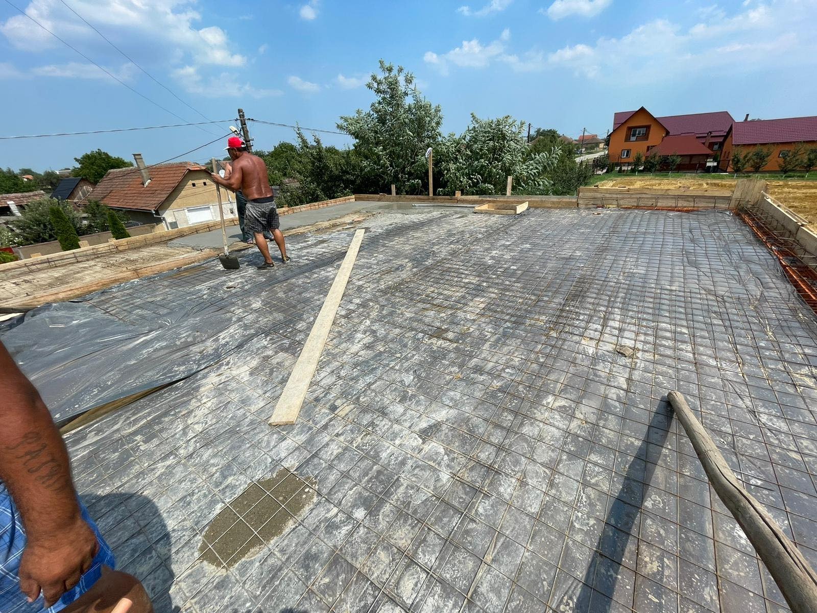 Przygotowanie dachu pod wylewkę betonową: zbrojenie siatką, folia izolacyjna. Widoczny dekarz z łopatą w czerwonym kasku. Prace dekarskie w słoneczny dzień.
