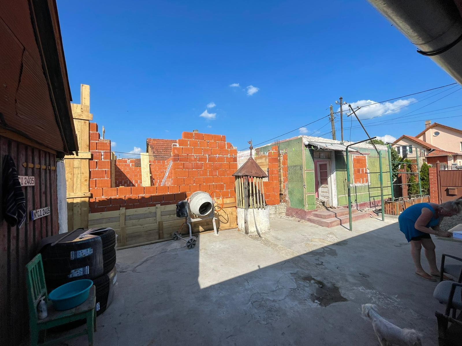 Budowa domu z czerwonej cegły w toku, widoczna betoniarka, studnia i kobieta w niebieskiej sukience. Częściowo wykończony dach sąsiedniego budynku.