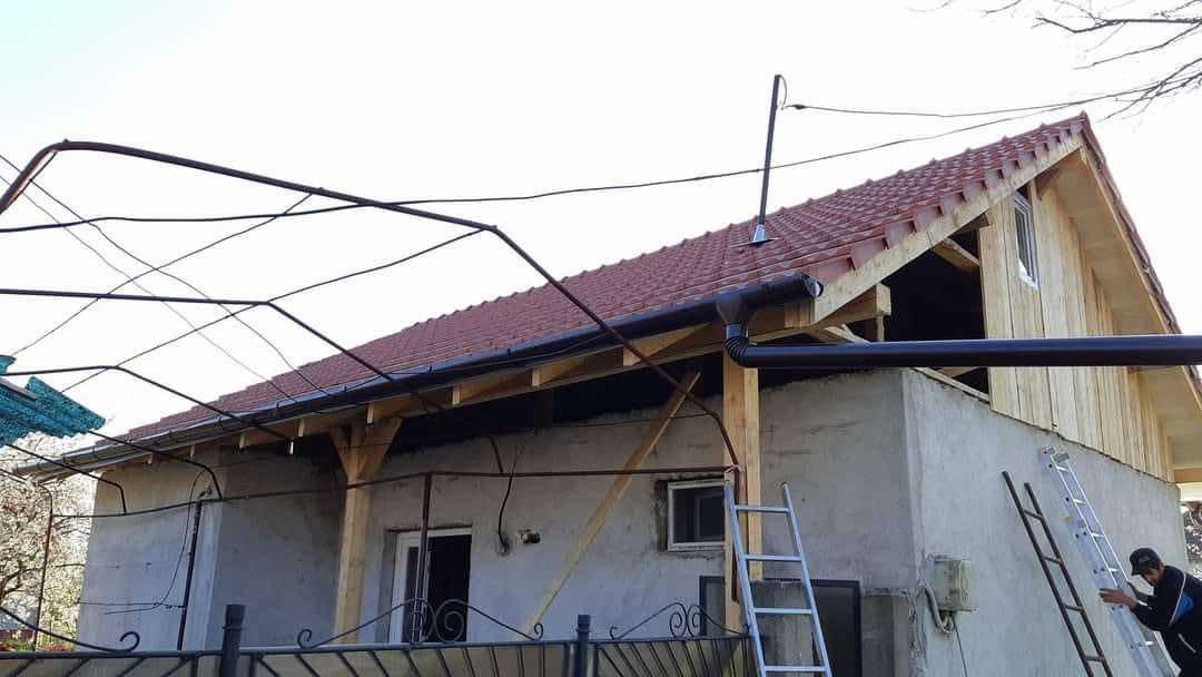 Dom w trakcie remontu dachu z nowymi rynnami, widoczne drewniane elementy konstrukcyjne i drabiny, pracownik na drabinie. Ujęcie z dołu, zza ozdobnego ogrodzenia.