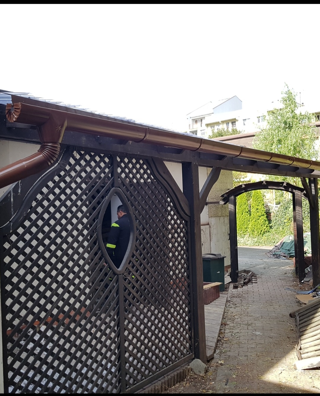 Brązowa rynna zamontowana na drewnianej konstrukcji z kratownicą, przez którą widać postać w uniformie. Widoczny fragment chodnika i zieleń w tle.