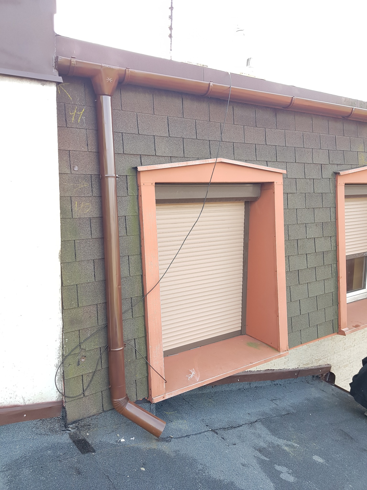 Brązowa rynna spustowa zamontowana na dachu z oknami w Katowicach. Widoczna elewacja budynku pokryta szarą dachówką bitumiczną i fragment dachu pokrytego papą.