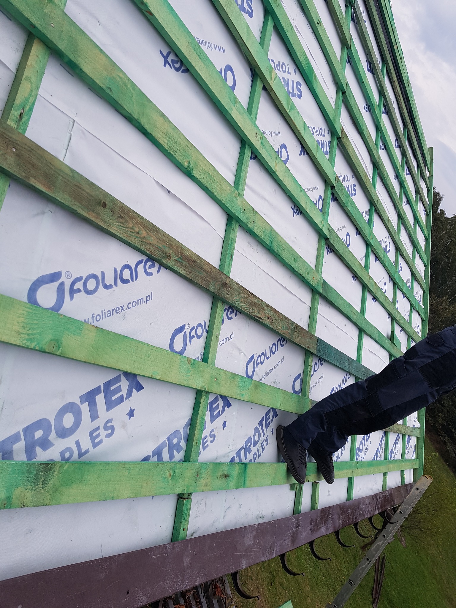 Montaż łat dachowych na membranie Foliarex i Trotex. Widoczne fragmenty ubrania montera. Całość z perspektywy od dołu, niebo w tle.