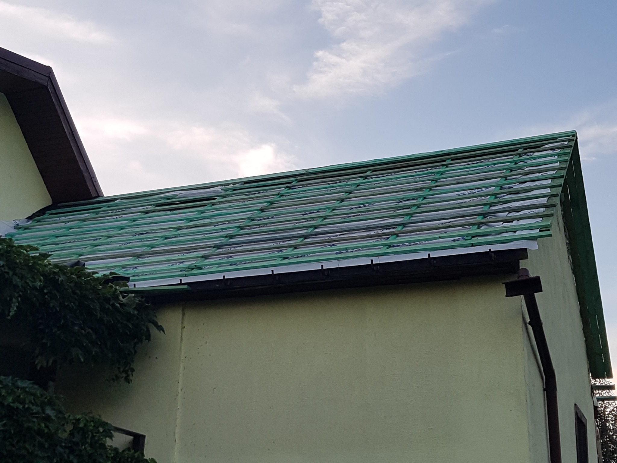 Dach budynku w trakcie wymiany, widoczne zielone łaty i membrana dachowa, ujęcie z dołu, fragment rynny i elewacja budynku w tle.