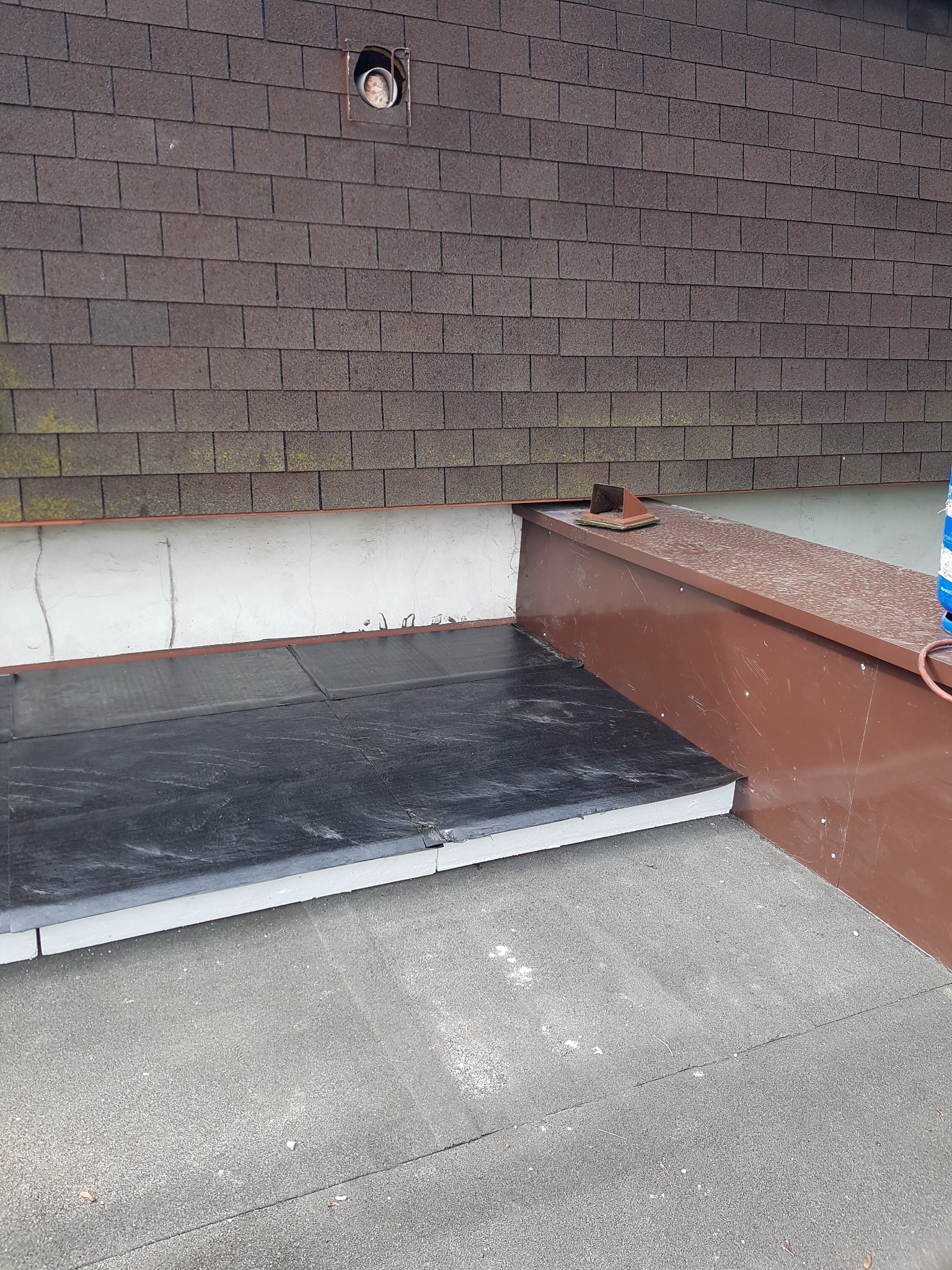 Fragment dachu z brązową obróbką blacharską i czarną izolacją. Widoczny komin wentylacyjny w brązowej elewacji z gontu bitumicznego.